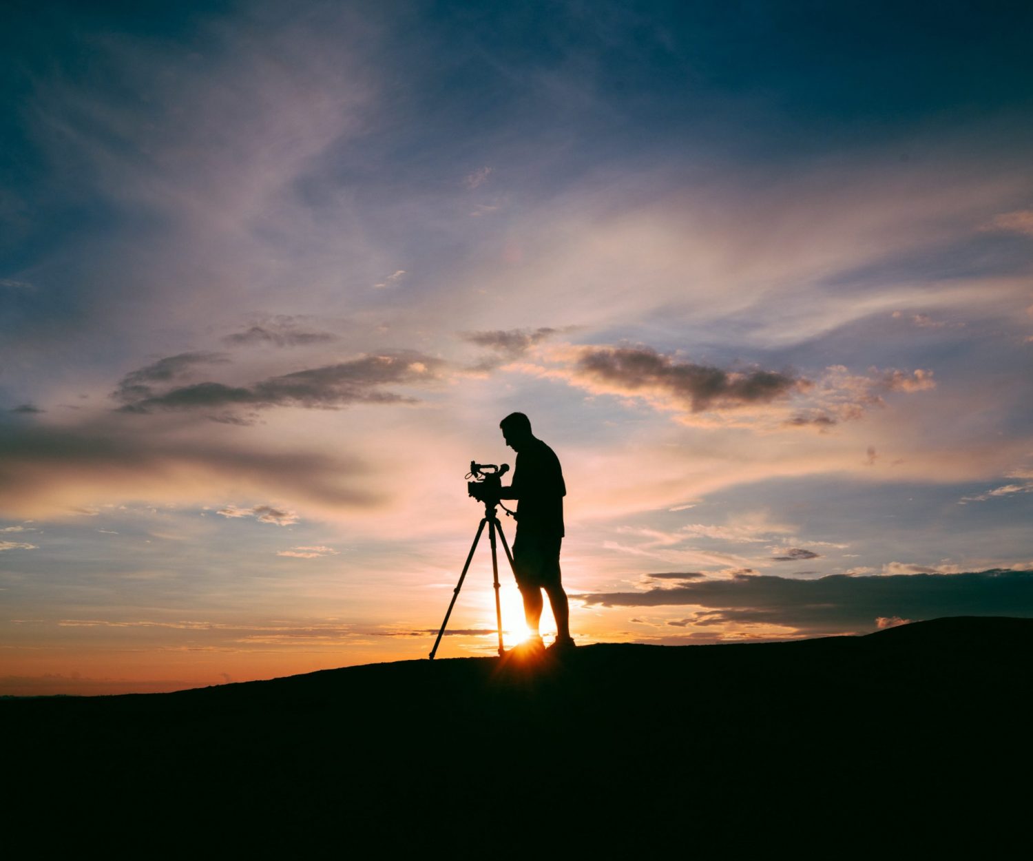 silhouette of man standing beside camera tripod during sunset
